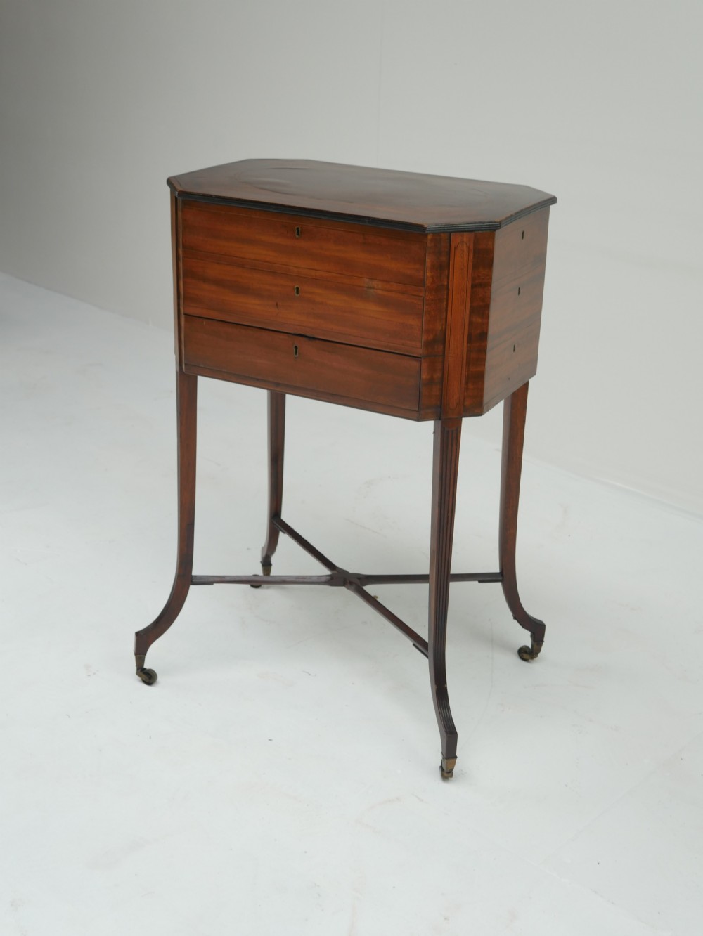 A Sheraton period mahogany occasional table featuring three stacked drawers, canted corners, splayed legs with brass castors, and an X-stretcher.