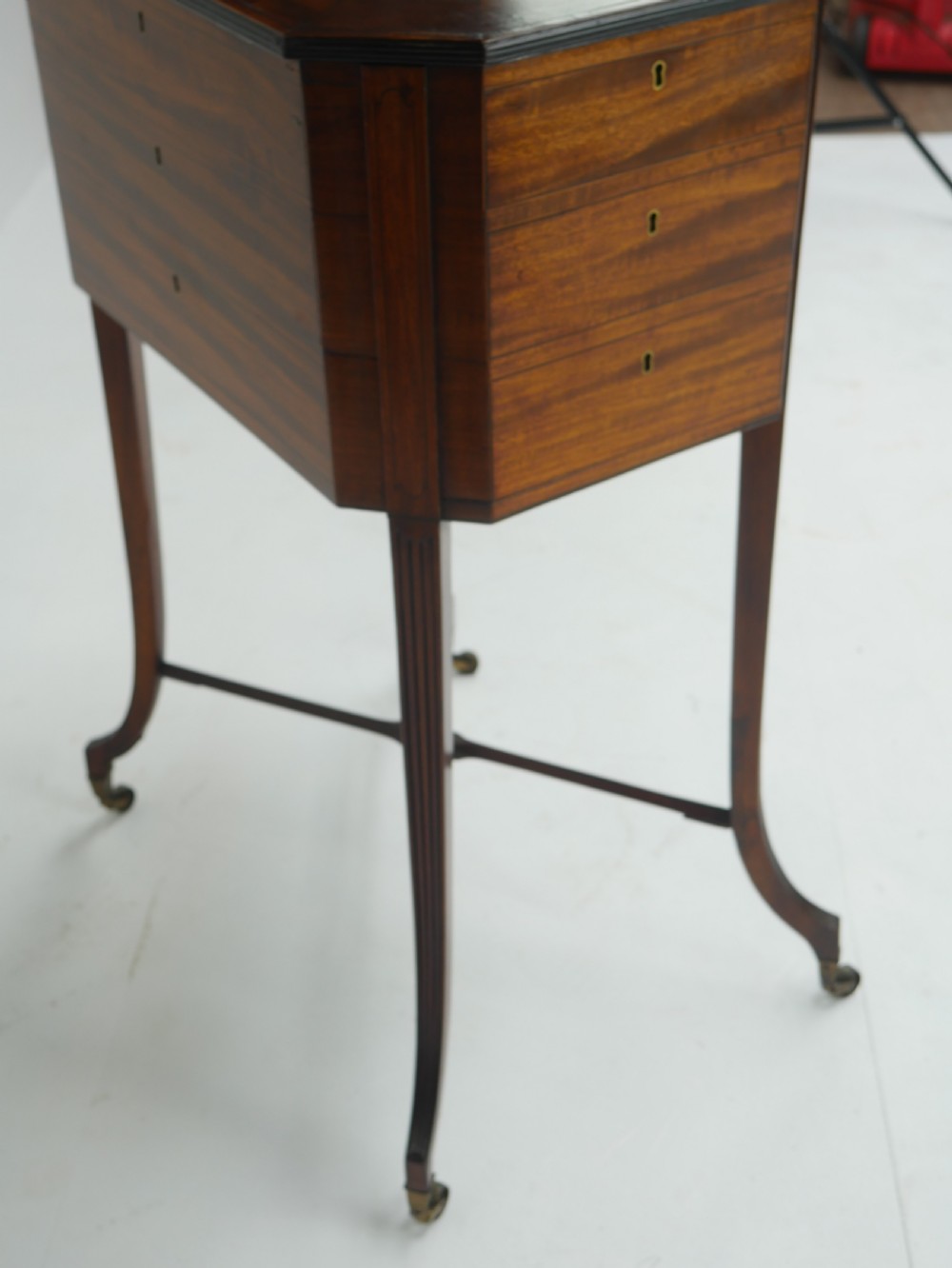 A Sheraton period mahogany occasional table featuring three stacked drawers, canted corners, splayed legs with brass castors, and an X-stretcher. View X
