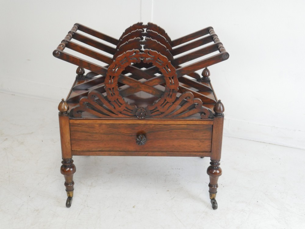 A Regency period dark rosewood music canterbury featuring a carved central wreath handle, slatted divided compartments, a single base drawer, and turned legs on brass castors.