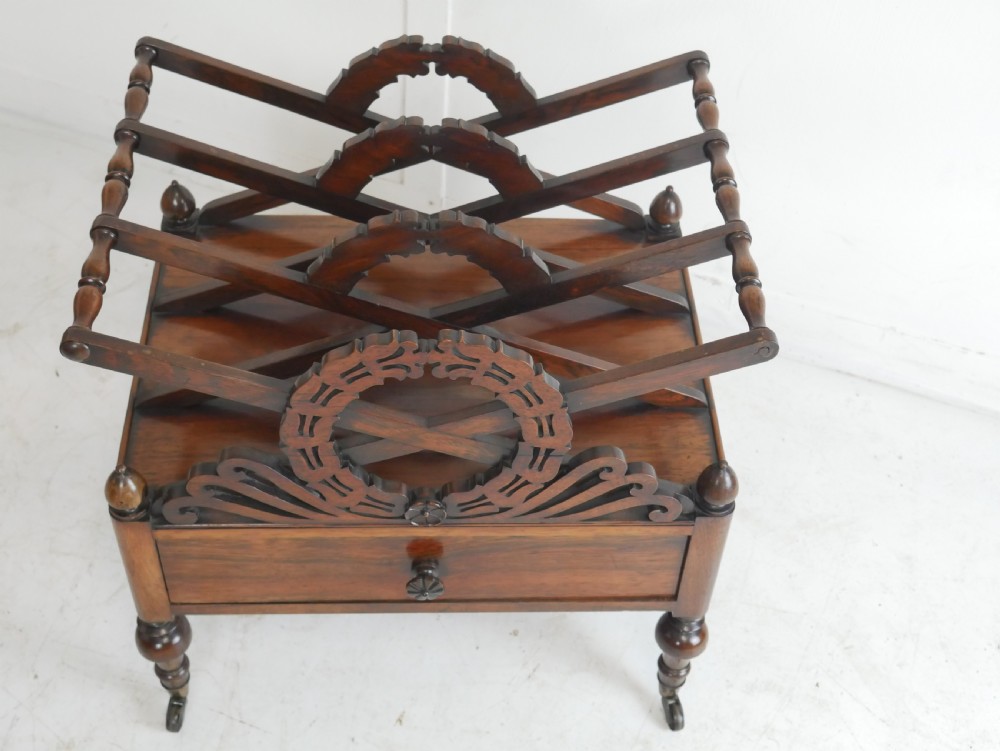 A Regency period dark rosewood music canterbury featuring a carved central wreath handle, slatted divided compartments, a single base drawer, and turned legs on brass castors. View II
