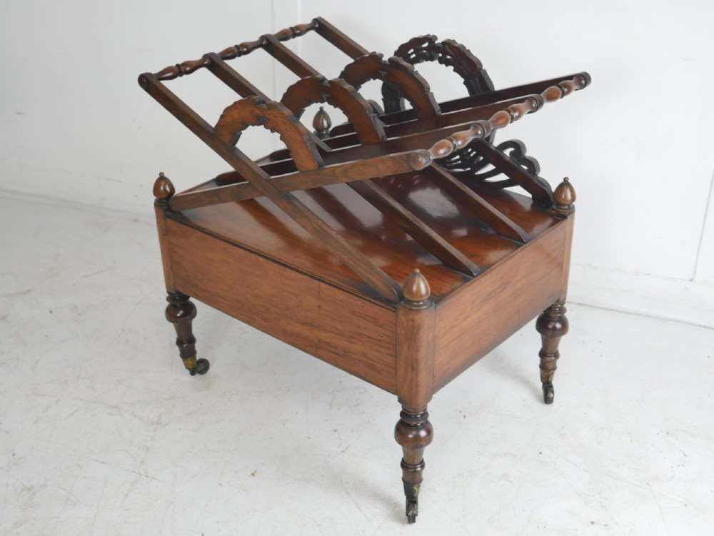A Regency period dark rosewood music canterbury featuring a carved central wreath handle, slatted divided compartments, a single base drawer, and turned legs on brass castors. View IV