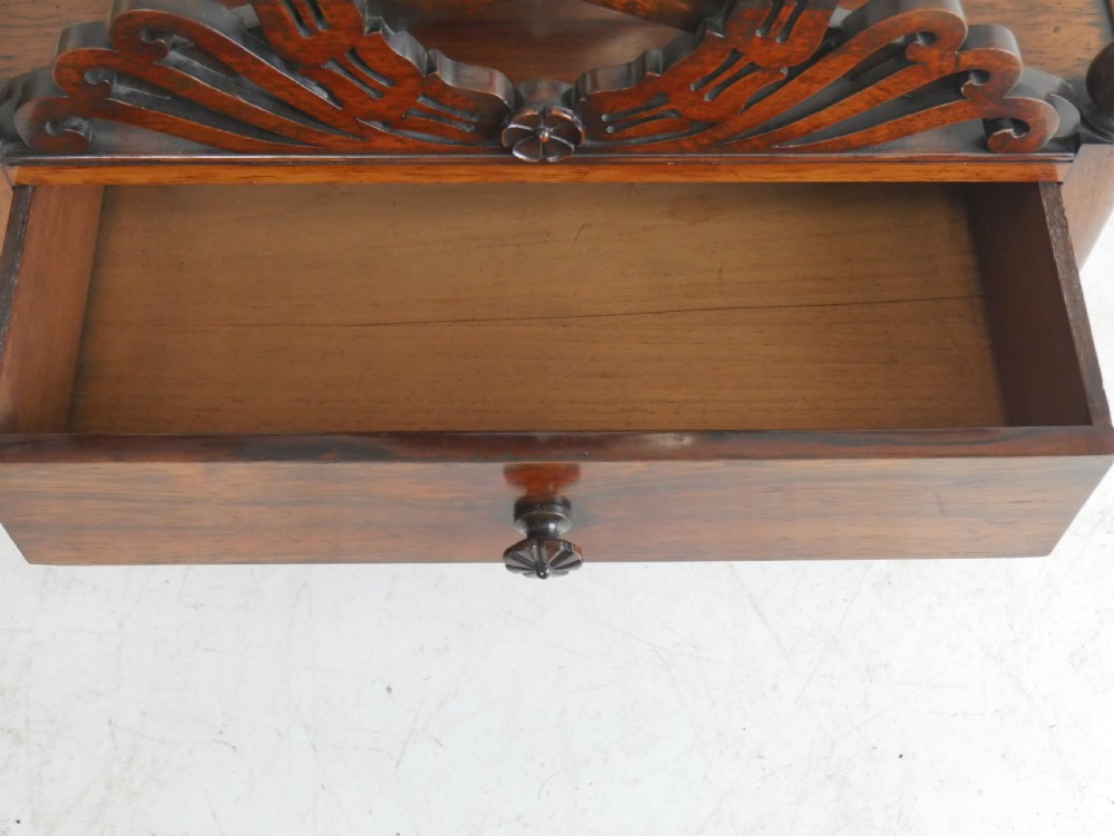 A Regency period dark rosewood music canterbury featuring a carved central wreath handle, slatted divided compartments, a single base drawer, and turned legs on brass castors. View V