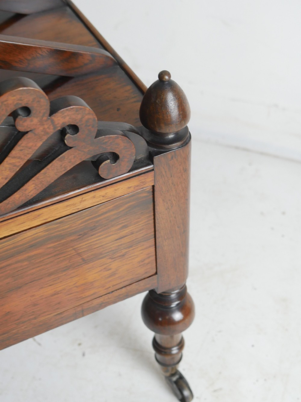 A Regency period dark rosewood music canterbury featuring a carved central wreath handle, slatted divided compartments, a single base drawer, and turned legs on brass castors. View VII