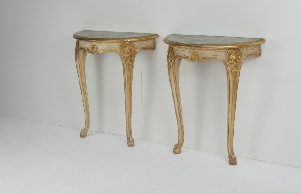 A pair of 1920s demi-lune wall console tables featuring cream and gilded finishes, carved cabriole legs, and dark tops.