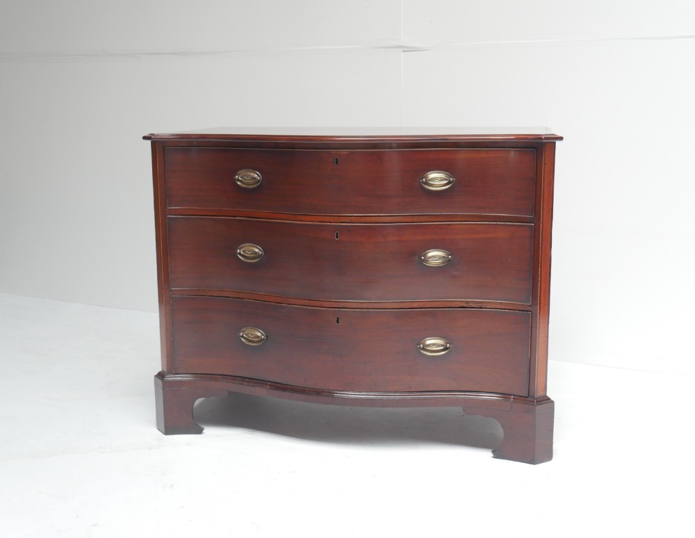 A Georgian dark mahogany chest of drawers with a serpentine curved front, three long drawers fitted with brass oval handles, standing on bracket feet. View XIV