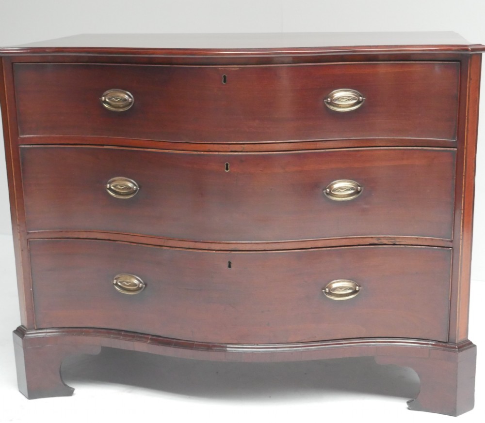 A Georgian dark mahogany chest of drawers with a serpentine curved front, three long drawers fitted with brass oval handles, standing on bracket feet. View X