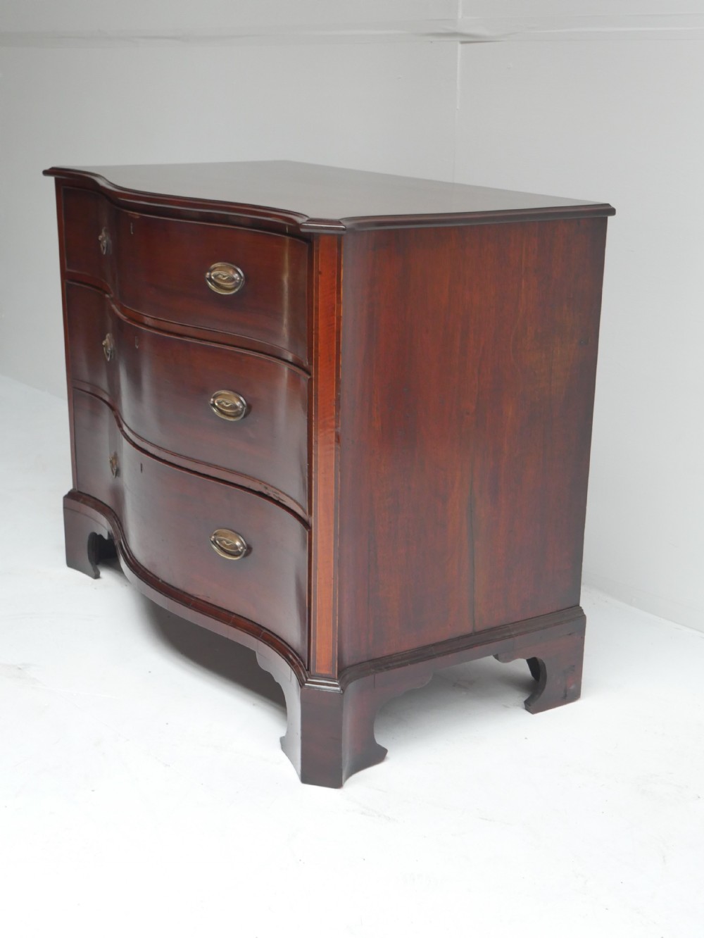 A Georgian dark mahogany chest of drawers with a serpentine curved front, three long drawers fitted with brass oval handles, standing on bracket feet. View II