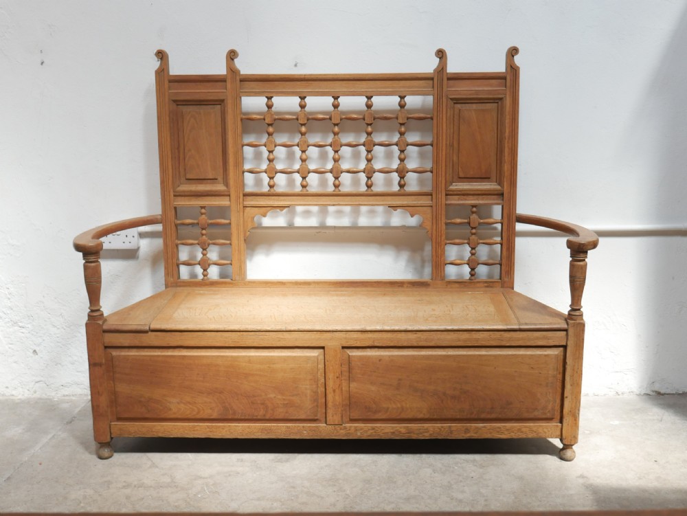 An antique light oak hall settle featuring a turned spindle lattice backrest, sweeping curved armrests, and a lift-up storage seat box.