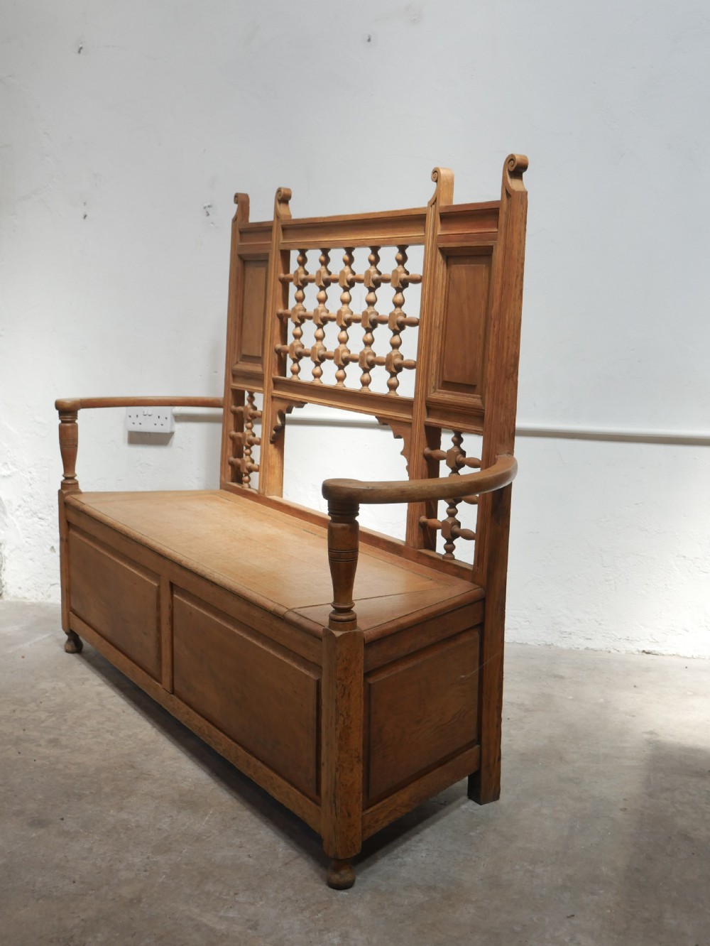 An antique light oak hall settle featuring a turned spindle lattice backrest, sweeping curved armrests, and a lift-up storage seat box. View II