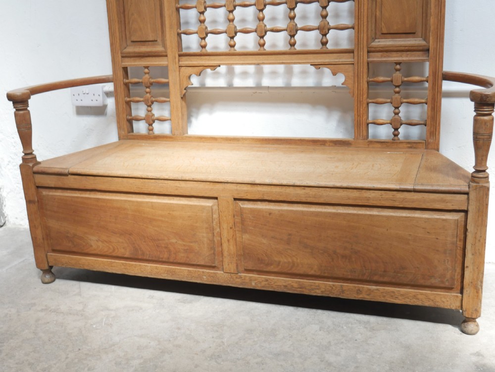 An antique light oak hall settle featuring a turned spindle lattice backrest, sweeping curved armrests, and a lift-up storage seat box. View X