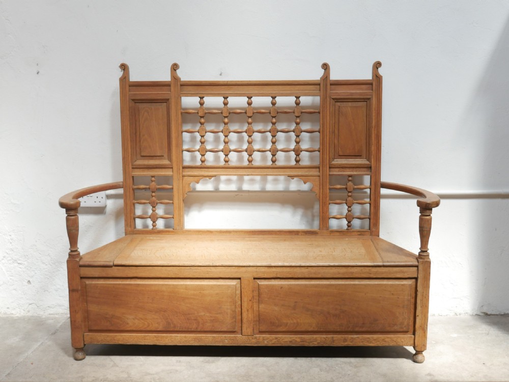 An antique light oak hall settle featuring a turned spindle lattice backrest, sweeping curved armrests, and a lift-up storage seat box. View XIV