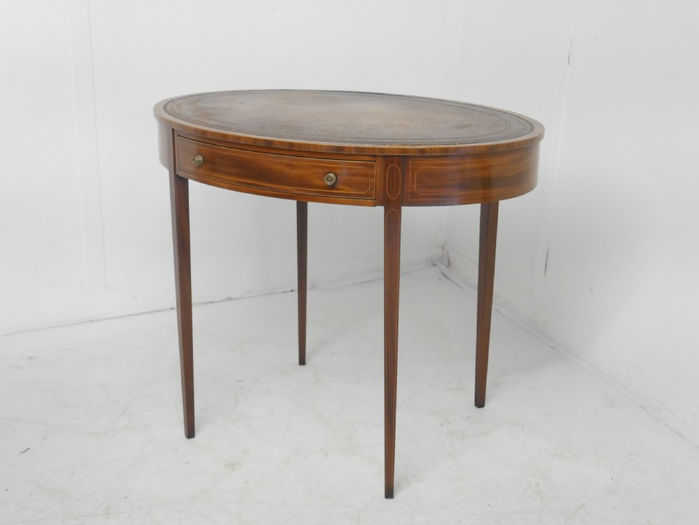 An oval mahogany centre table featuring a brown leather inset top, a single drawer with twin brass knobs, satinwood string inlay, and four square tapered legs.