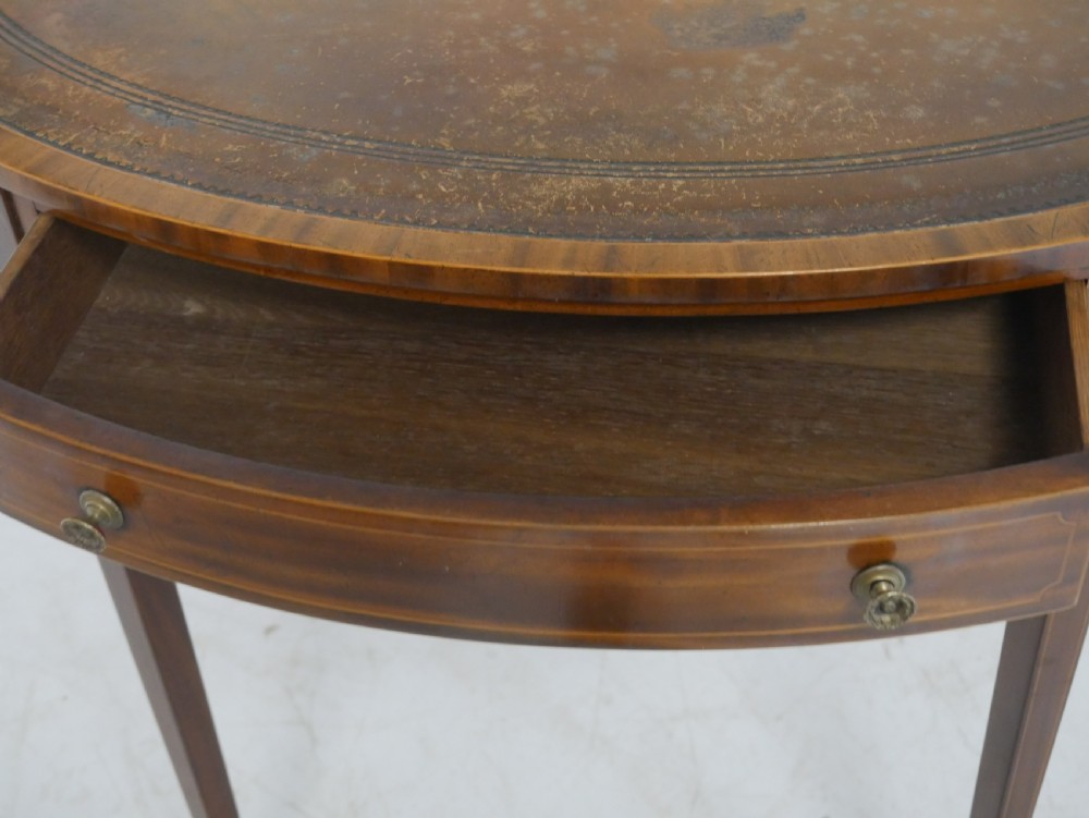An oval mahogany centre table featuring a brown leather inset top, a single drawer with twin brass knobs, satinwood string inlay, and four square tapered legs. View VI