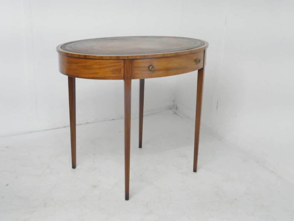 An oval mahogany centre table featuring a brown leather inset top, a single drawer with twin brass knobs, satinwood string inlay, and four square tapered legs. View XI