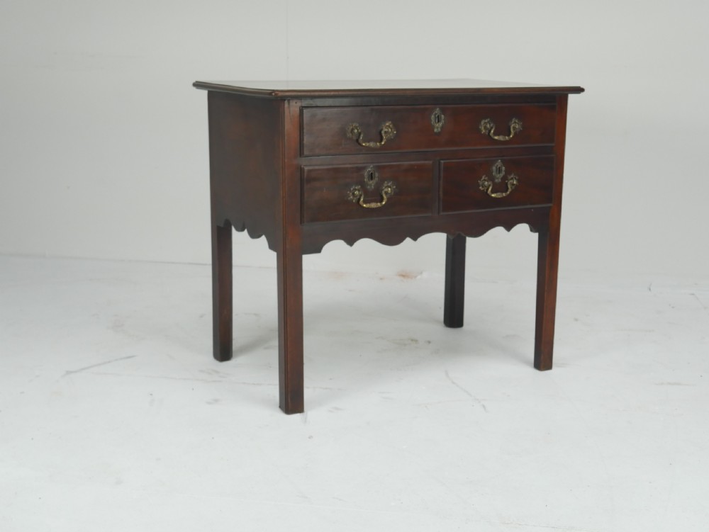 A dark brown Georgian mahogany lowboy featuring three drawers with brass handles and a shaped lower apron on square legs.