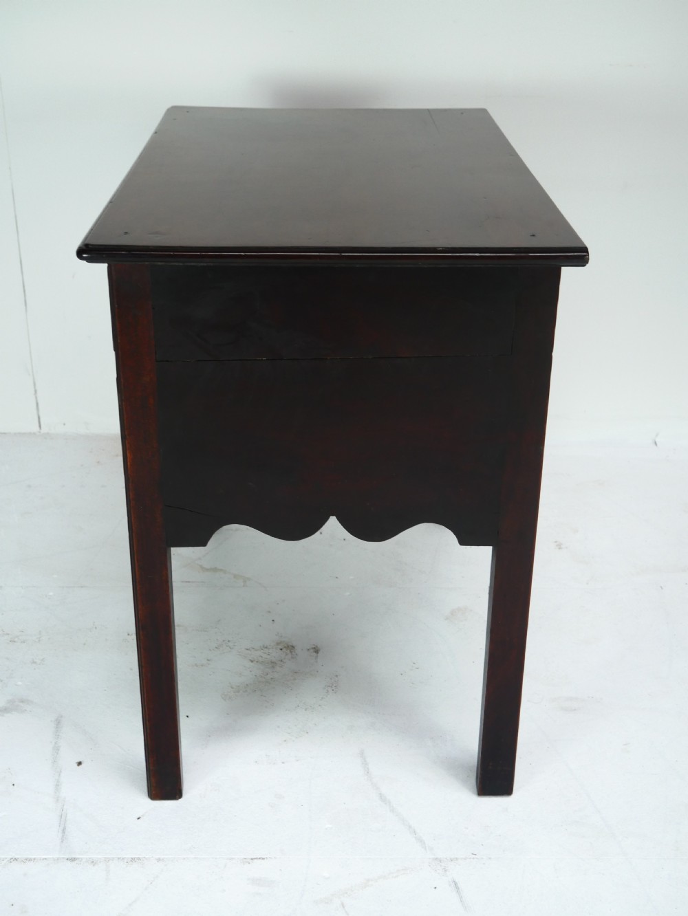 A dark brown Georgian mahogany lowboy featuring three drawers with brass handles and a shaped lower apron on square legs. View XI