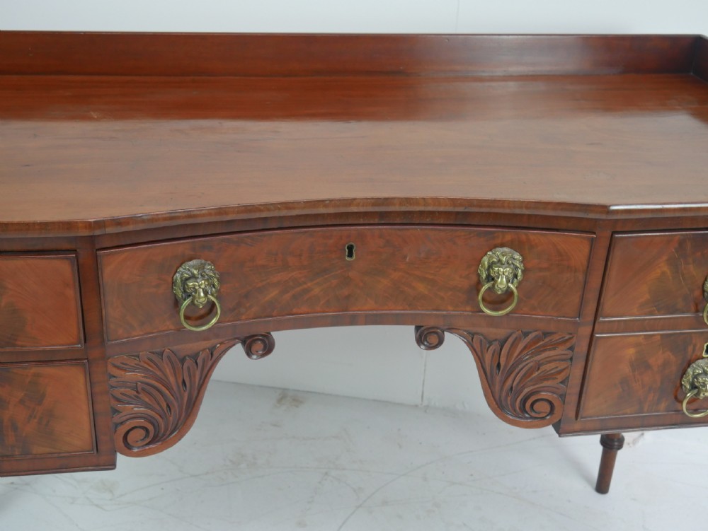 A Regency flame mahogany sideboard featuring five drawers with brass lion mask handles, turned reeded legs, and carved corner brackets. View II
