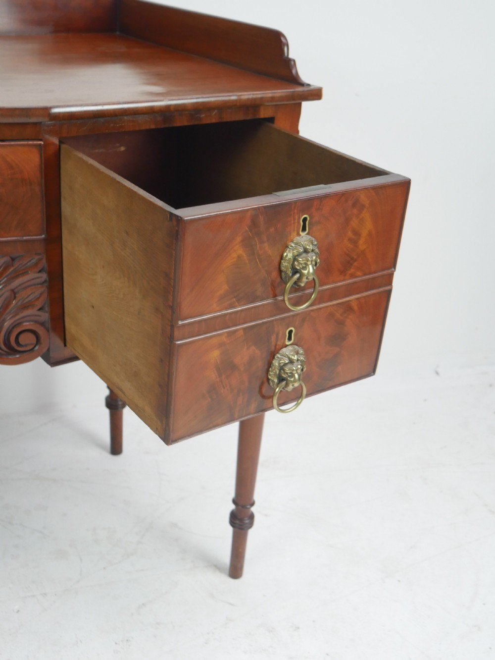 A Regency flame mahogany sideboard featuring five drawers with brass lion mask handles, turned reeded legs, and carved corner brackets. View VI