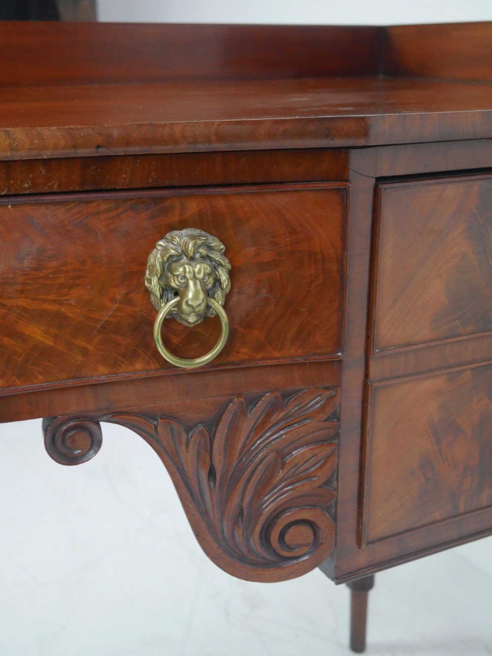 A Regency flame mahogany sideboard featuring five drawers with brass lion mask handles, turned reeded legs, and carved corner brackets. View XV