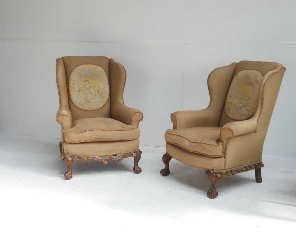 A pair of 1920s wingback armchairs upholstered in gold fabric featuring oval embroidered back panels and carved walnut ball and claw feet.