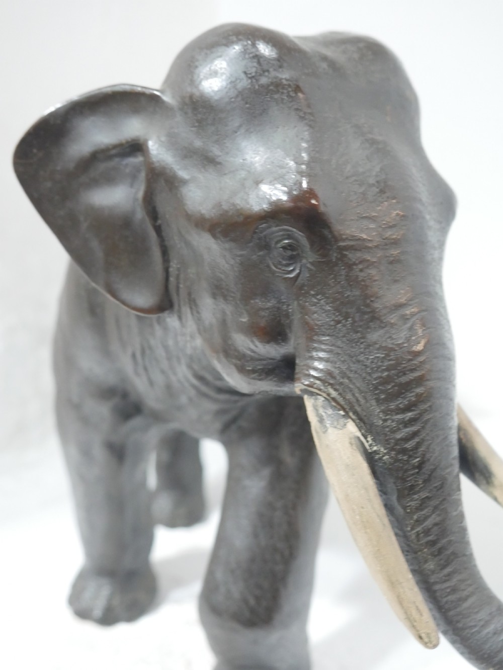 A dark patinated bronze figure of an elephant in mid-stride featuring light-coloured tusks and textured skin detail. View VI