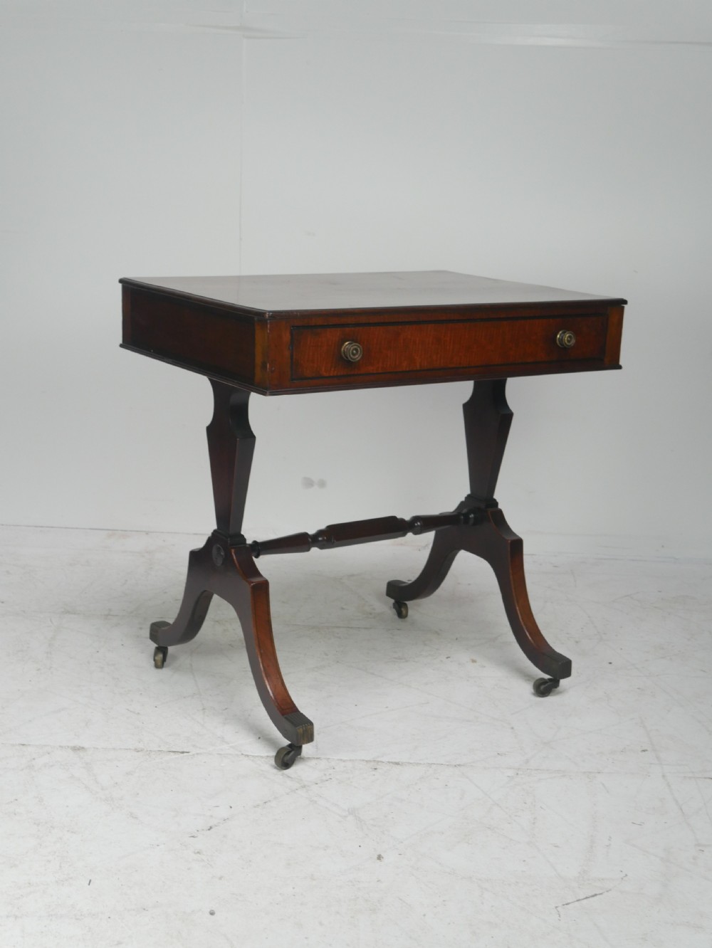 A Regency mahogany side table featuring a single drawer with brass knobs, a turned cross stretcher, and splayed legs on castors.