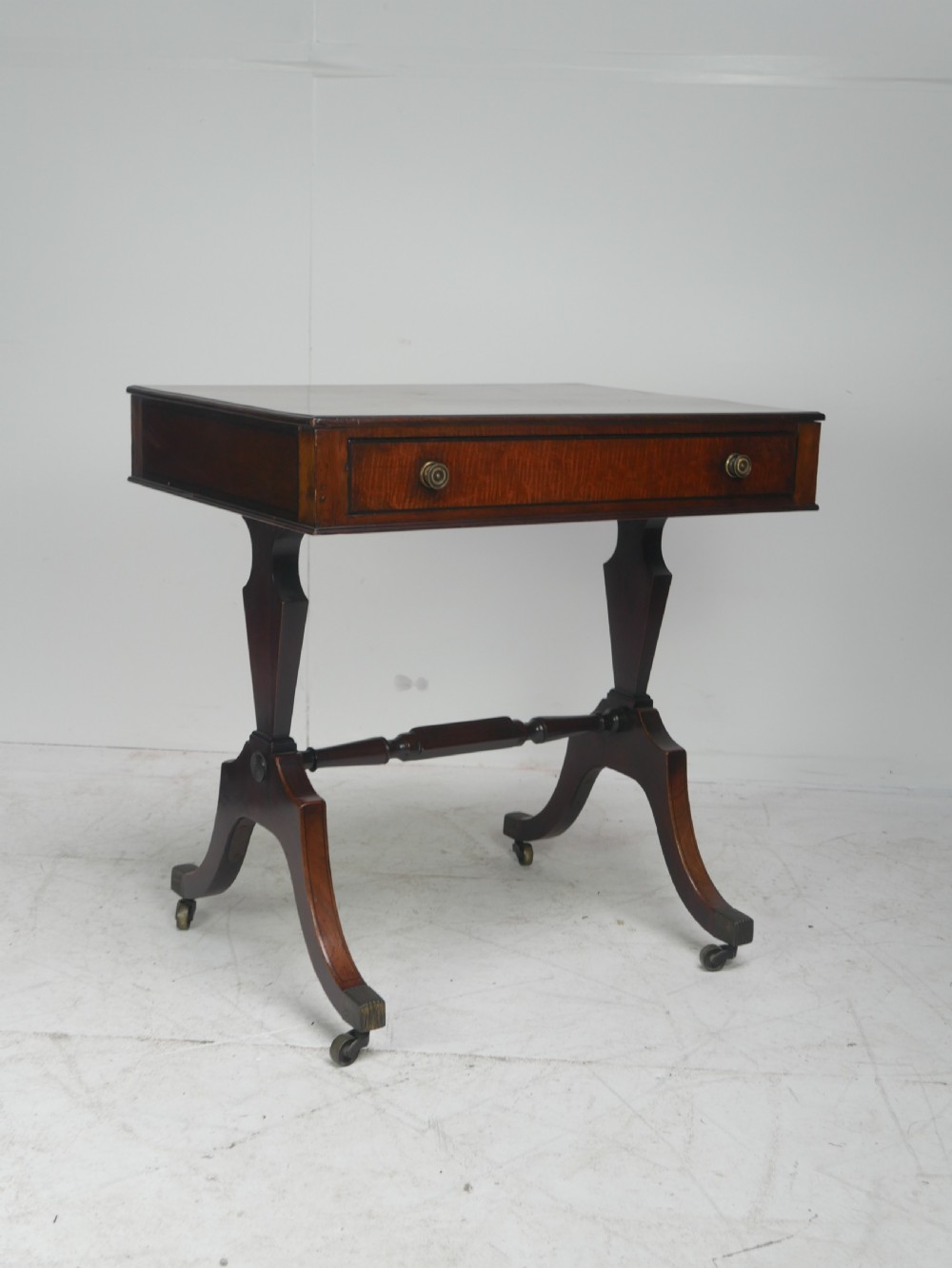 A Regency mahogany side table featuring a single drawer with brass knobs, a turned cross stretcher, and splayed legs on castors. View XI