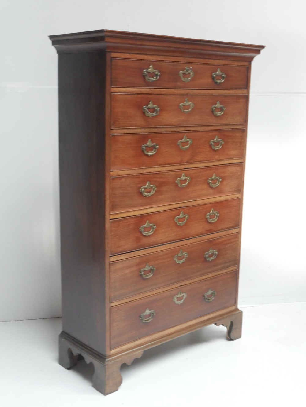 A tall, narrow mahogany chest of drawers featuring seven graduated drawers with brass drop handles and decorative shaped bracket feet.