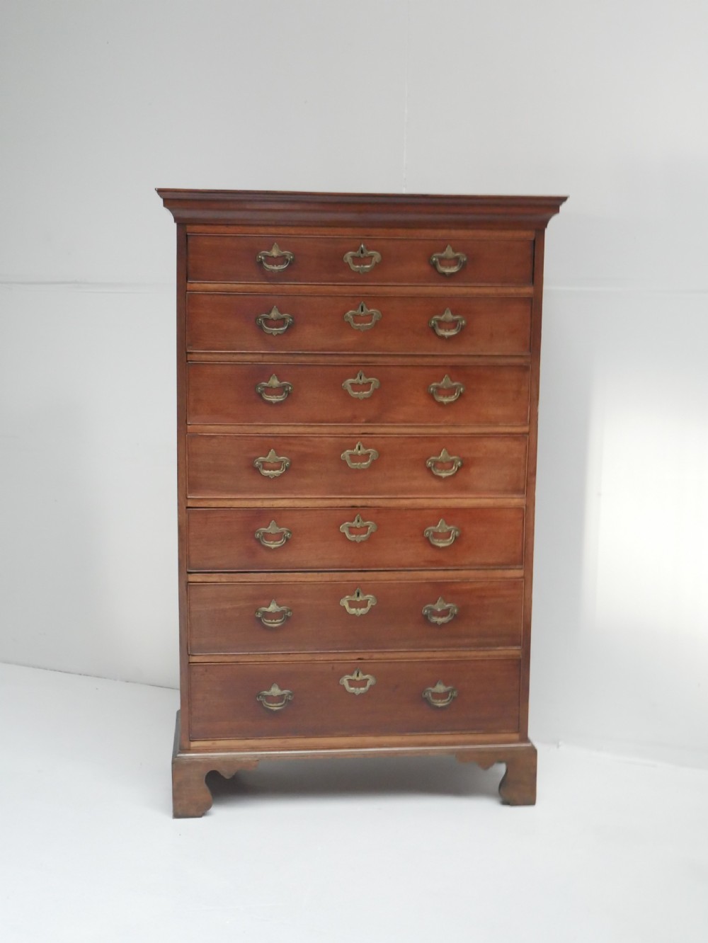 A tall, narrow mahogany chest of drawers featuring seven graduated drawers with brass drop handles and decorative shaped bracket feet. View III