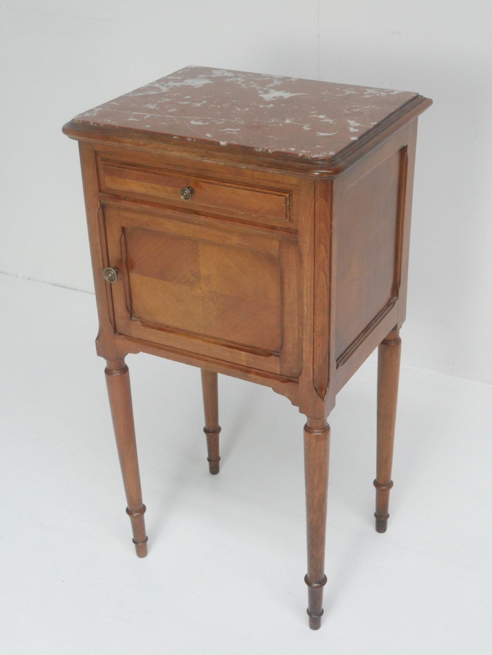 A French provincial walnut bedside cabinet with a red marble top, single drawer, inlaid wooden door, and slender turned legs.