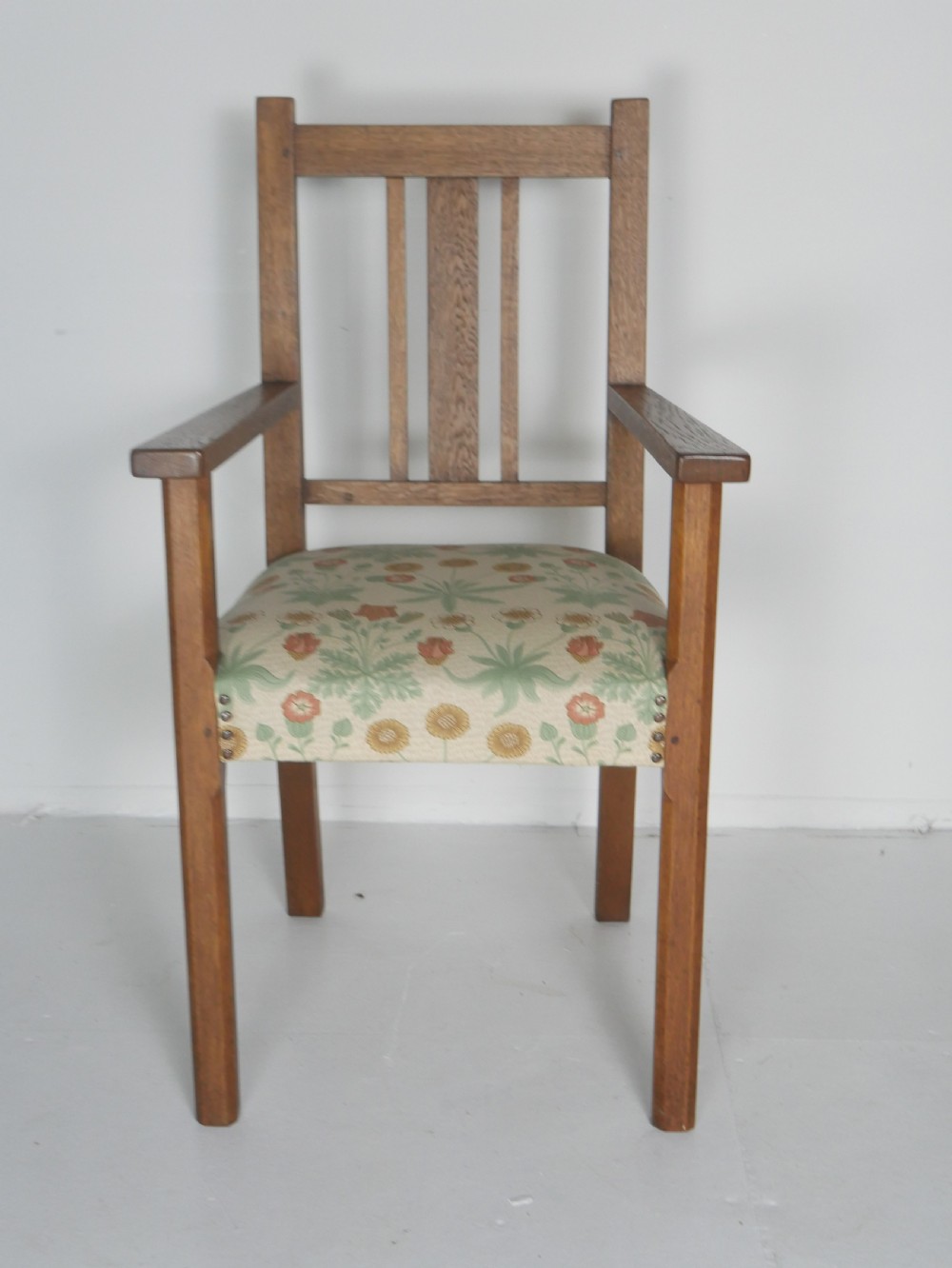 An Arts and Crafts solid oak armchair with a slatted backrest, flat armrests, and a botanical patterned fabric seat secured with studs.