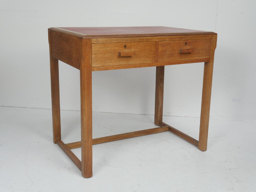 A light oak childs writing desk featuring a red inset leather top, two drawers with wooden pulls, and a square-section frame with a low floor stretcher.