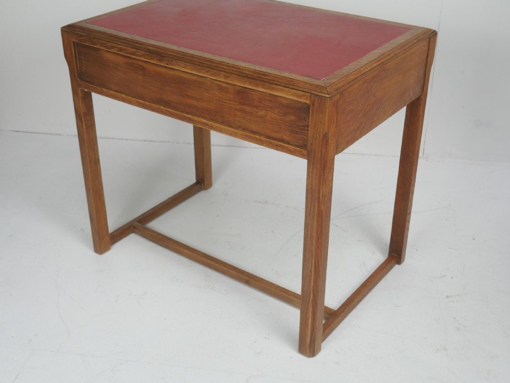 A light oak childs writing desk featuring a red inset leather top, two drawers with wooden pulls, and a square-section frame with a low floor stretcher. View VI