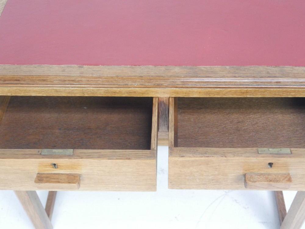 A light oak childs writing desk featuring a red inset leather top, two drawers with wooden pulls, and a square-section frame with a low floor stretcher. View XII