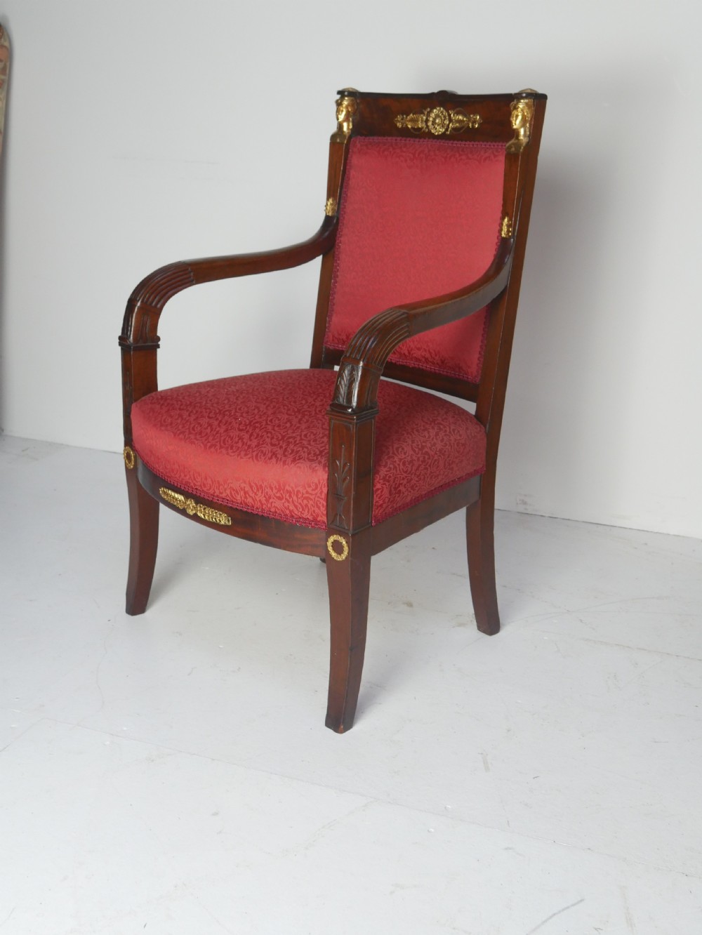A French Empire style mahogany armchair featuring red damask fabric upholstery, carved armrests, and decorative gilded bronze mounts.