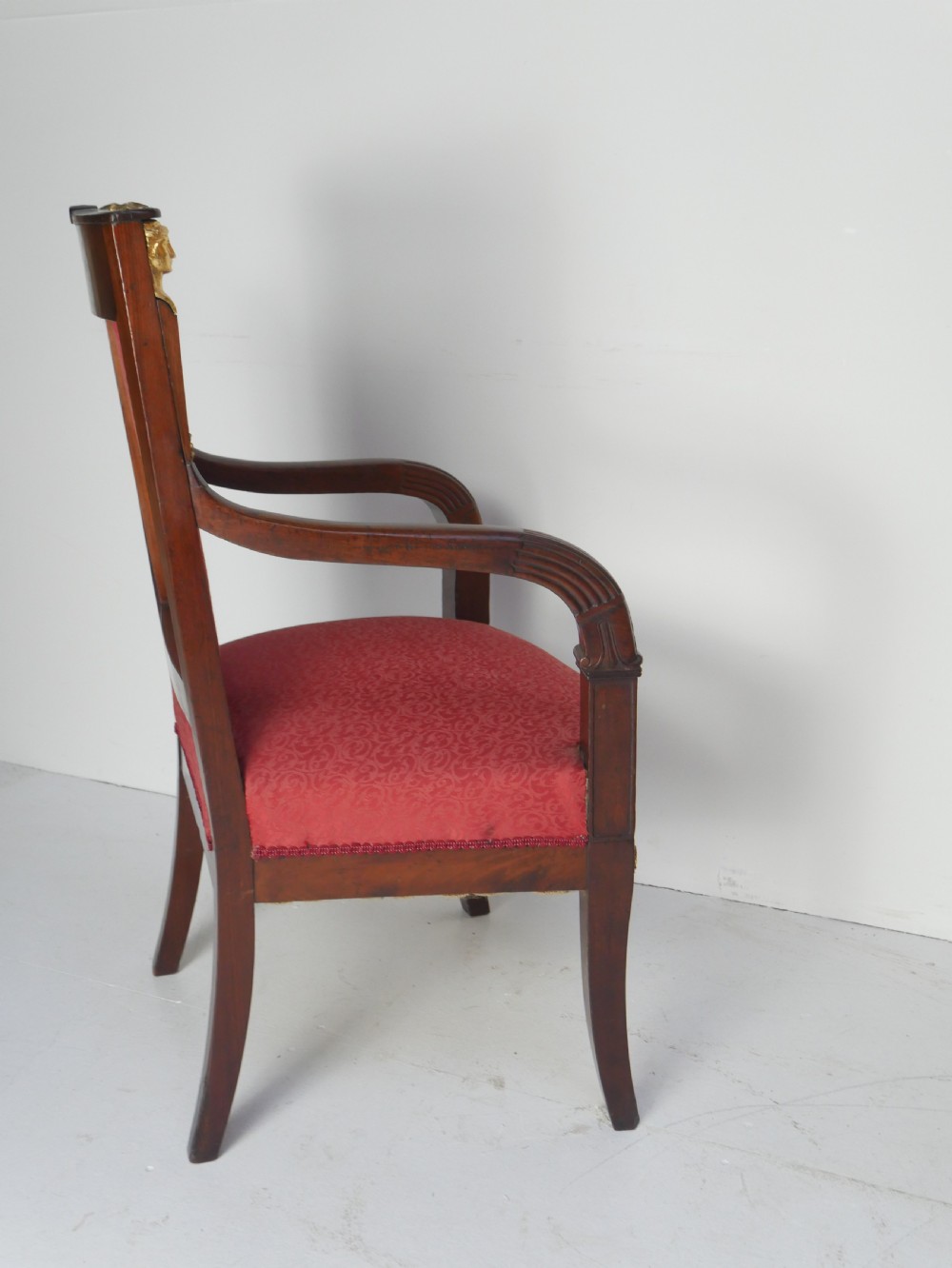 A French Empire style mahogany armchair featuring red damask fabric upholstery, carved armrests, and decorative gilded bronze mounts. View X
