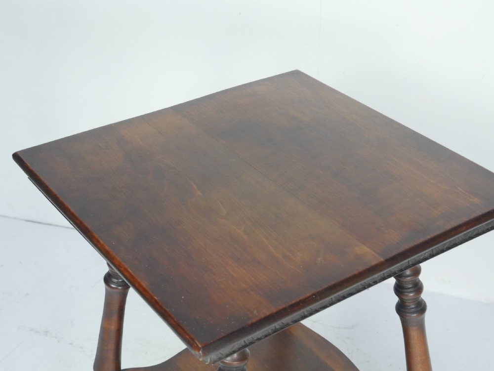 A dark brown wooden 1920s lamp table featuring four turned legs, a square top with carved edge detailing, and a lower tier shelf. View II