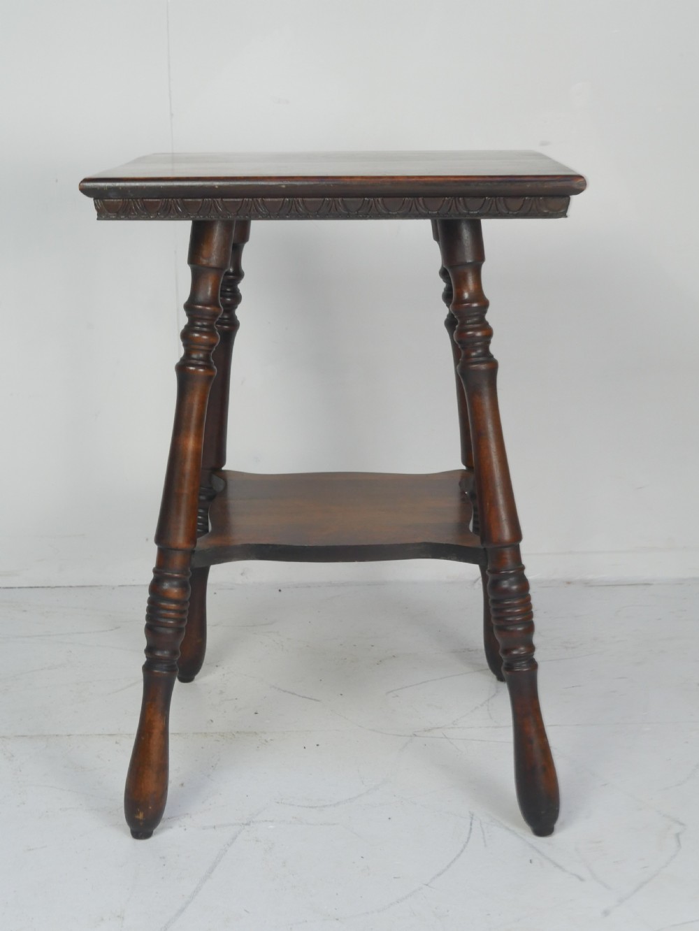 A dark brown wooden 1920s lamp table featuring four turned legs, a square top with carved edge detailing, and a lower tier shelf. View III