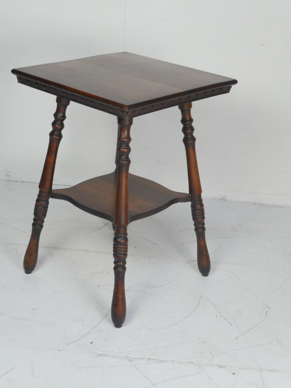 A dark brown wooden 1920s lamp table featuring four turned legs, a square top with carved edge detailing, and a lower tier shelf. View XI