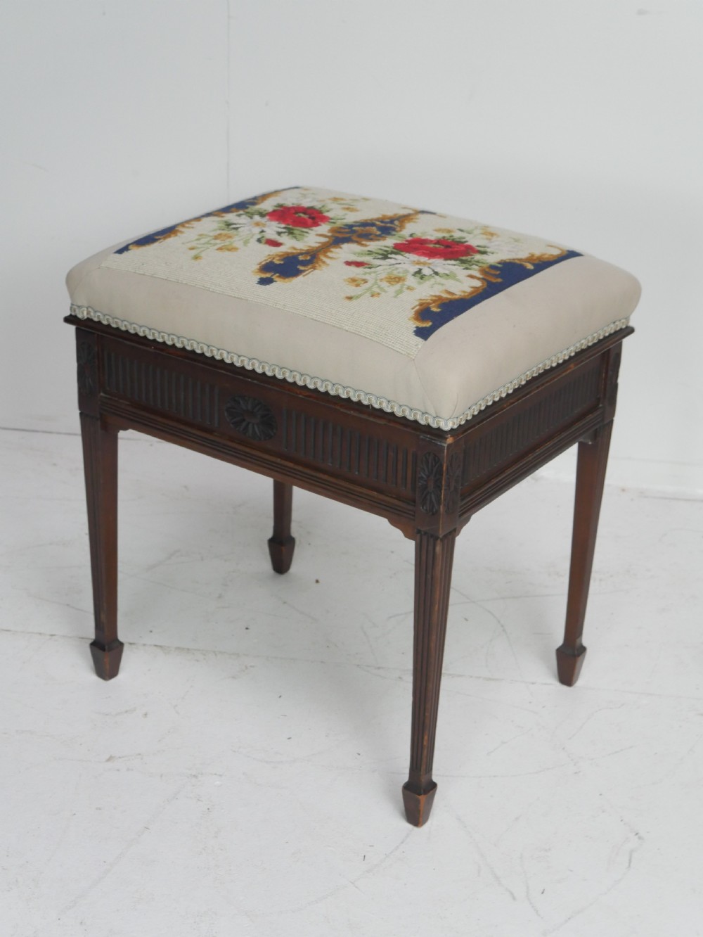 A rectangular antique mahogany piano stool featuring fluted tapered legs, carved central rosettes, and a floral needlepoint tapestry top.