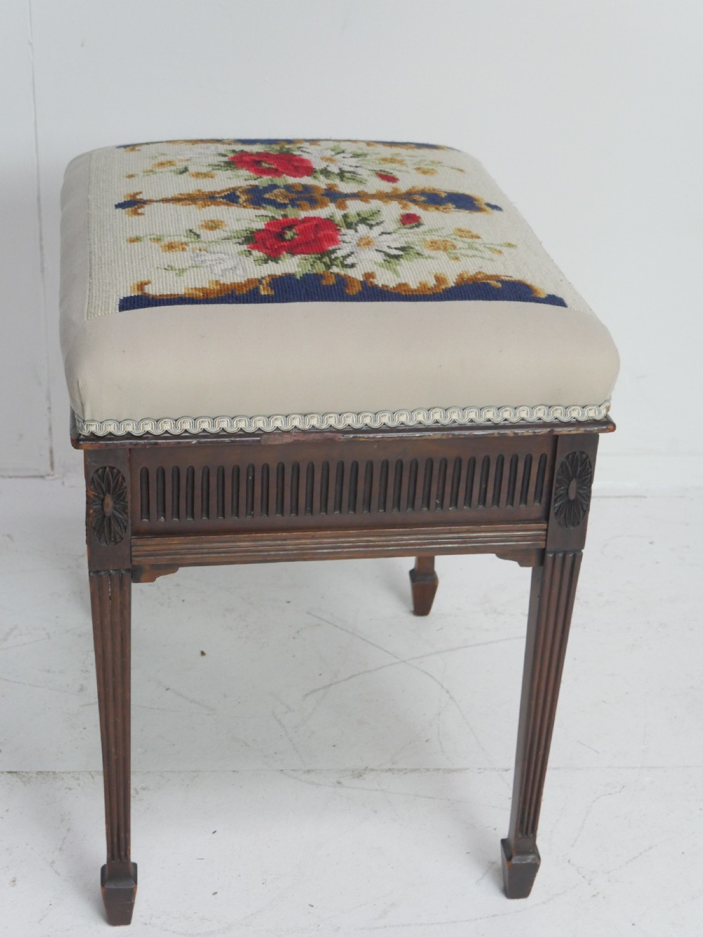 A rectangular antique mahogany piano stool featuring fluted tapered legs, carved central rosettes, and a floral needlepoint tapestry top. View V