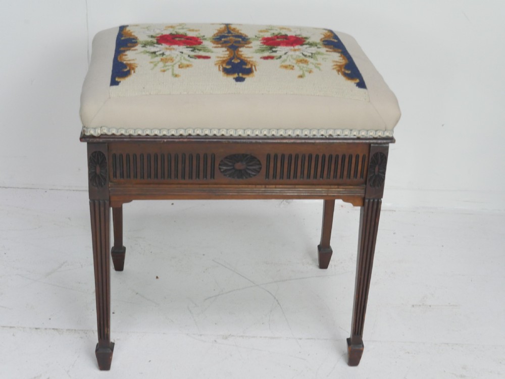 A rectangular antique mahogany piano stool featuring fluted tapered legs, carved central rosettes, and a floral needlepoint tapestry top. View VI