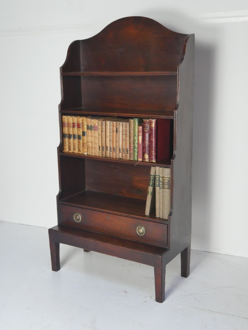 A dark mahogany Georgian waterfall bookcase featuring three open graduated shelves above a single drawer with brass handles.
