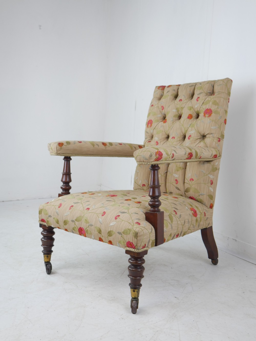 A 19th-century Victorian armchair featuring beige floral upholstery, a deep button-back rest, padded armrests, and turned mahogany legs set on brass casters.