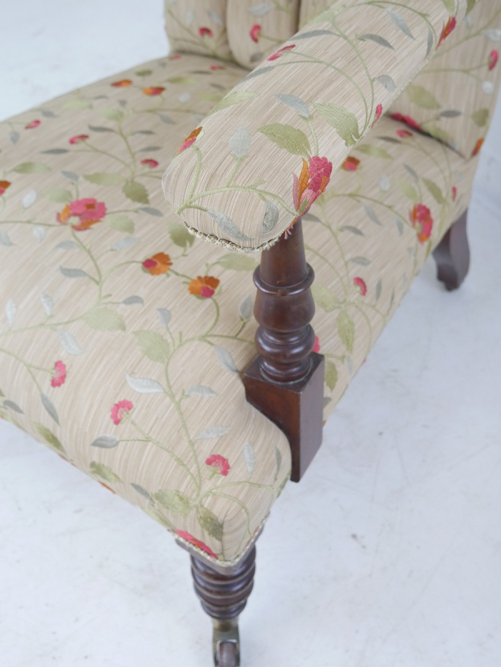 A 19th-century Victorian armchair featuring beige floral upholstery, a deep button-back rest, padded armrests, and turned mahogany legs set on brass casters. View V