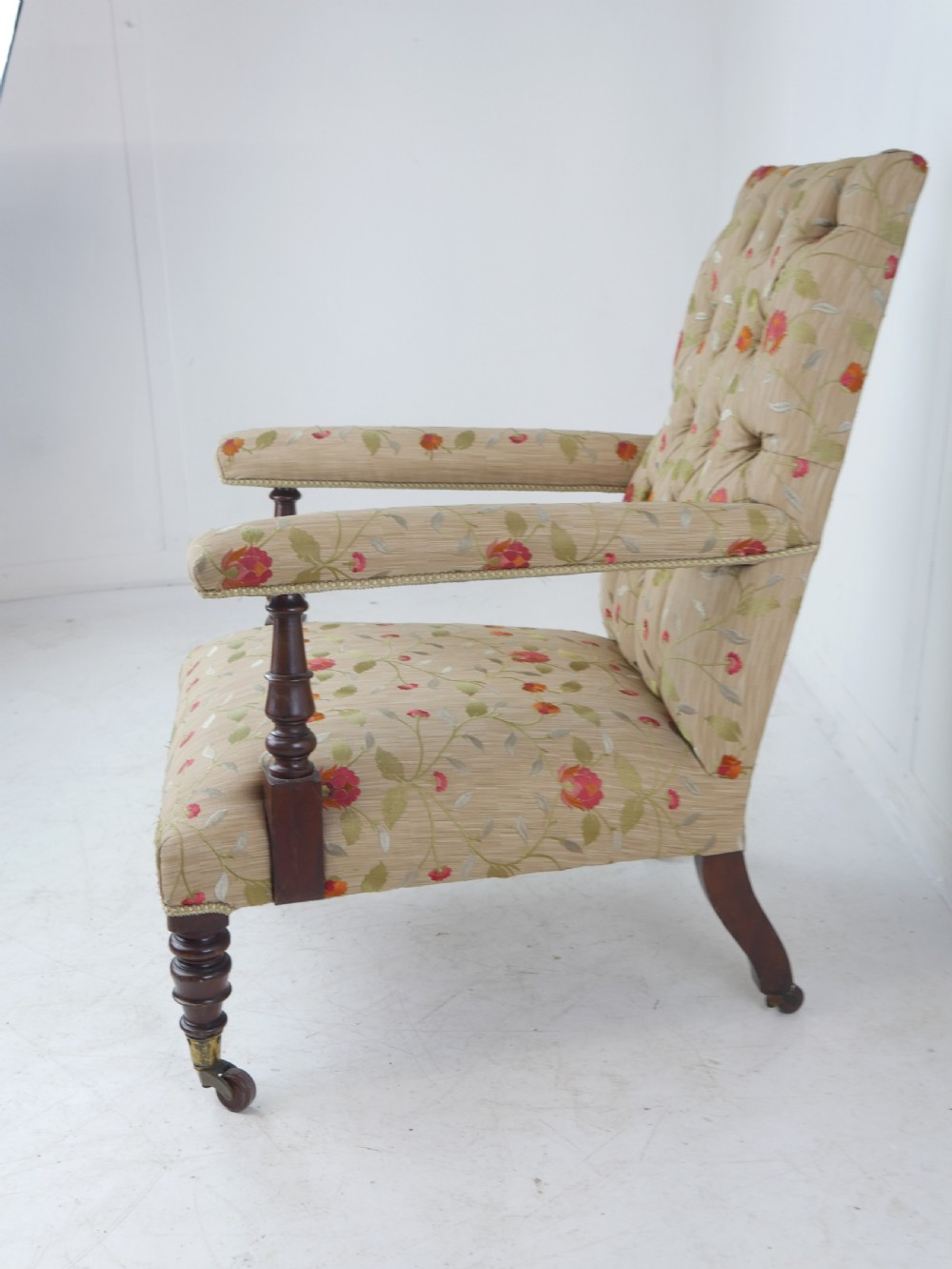 A 19th-century Victorian armchair featuring beige floral upholstery, a deep button-back rest, padded armrests, and turned mahogany legs set on brass casters. View VI
