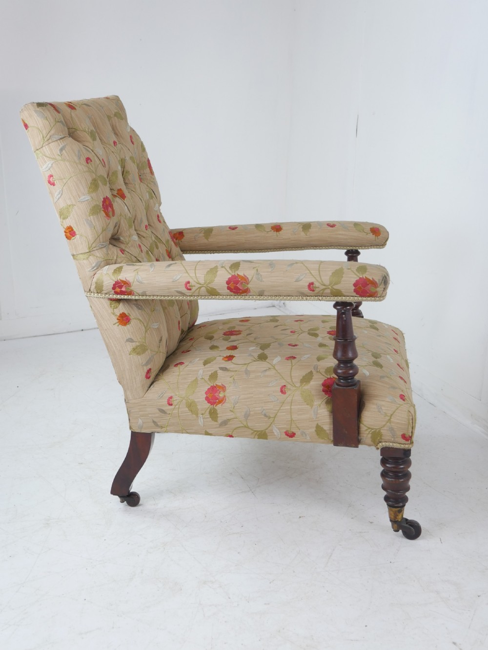 A 19th-century Victorian armchair featuring beige floral upholstery, a deep button-back rest, padded armrests, and turned mahogany legs set on brass casters. View X