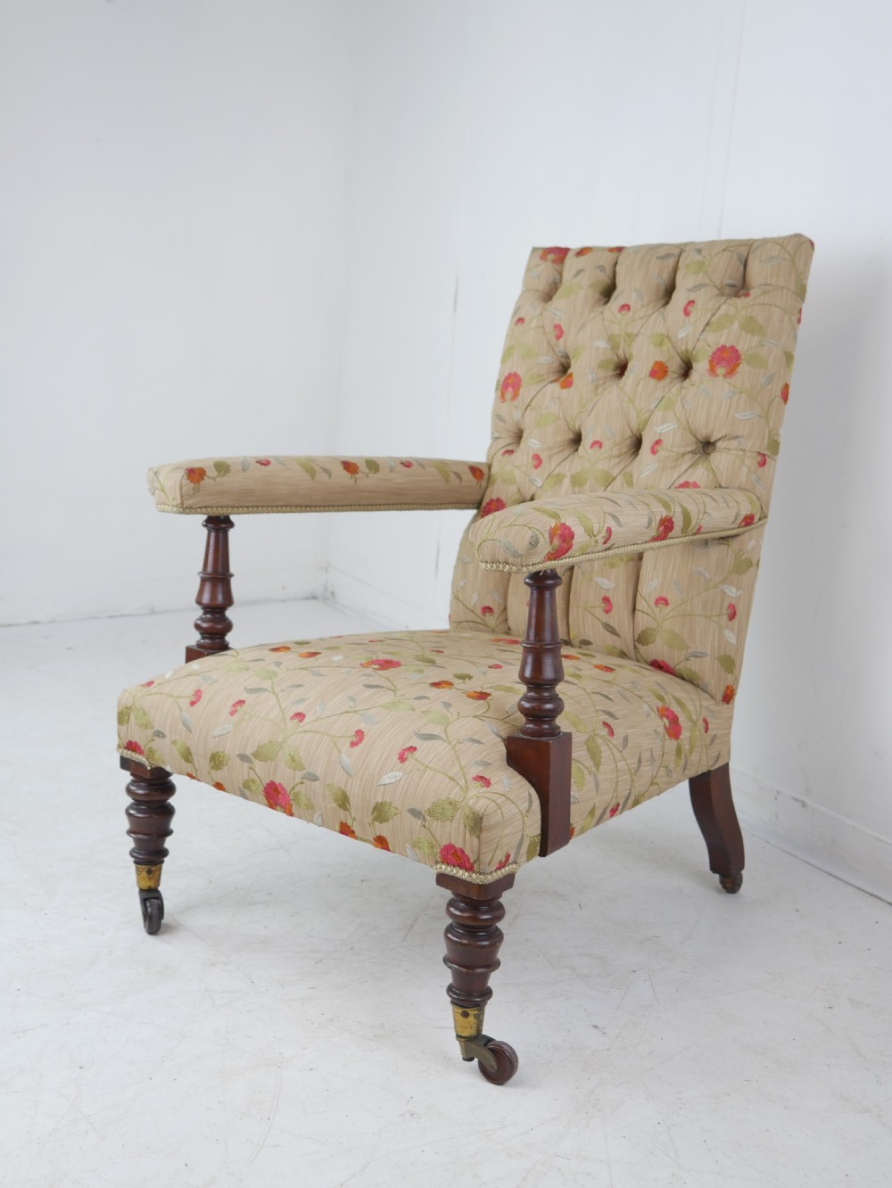 A 19th-century Victorian armchair featuring beige floral upholstery, a deep button-back rest, padded armrests, and turned mahogany legs set on brass casters. View XII