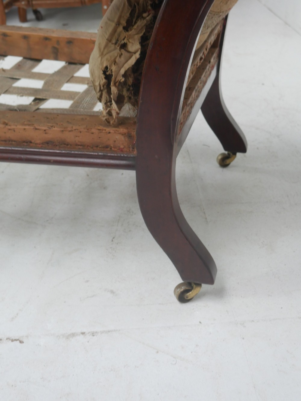 A 19th-century mahogany armchair frame featuring carved scroll arm terminals, turned front legs on brass castors, and worn calico lining ready for upholstery. View V