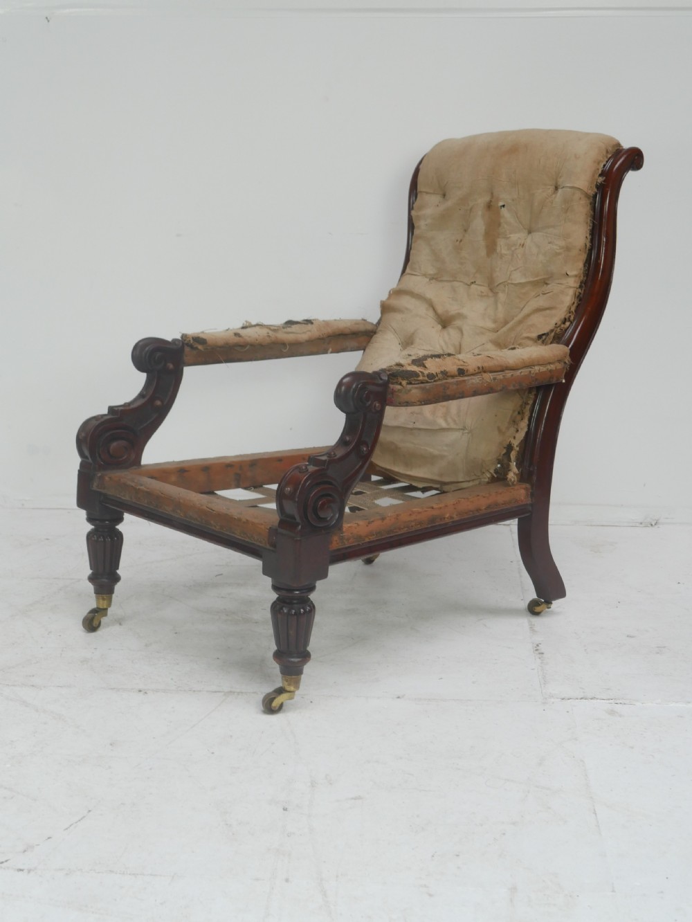 A 19th-century mahogany armchair frame featuring carved scroll arm terminals, turned front legs on brass castors, and worn calico lining ready for upholstery. View XII